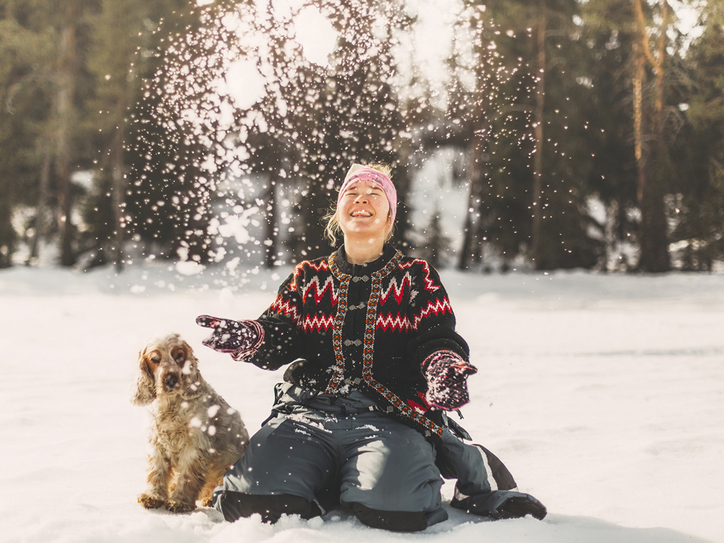 En ung kvinna sitter i snön med en hund bredvid sig. Hon ser glad ut och kastar snö som singlar ner på henne.