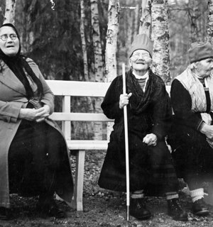 Fotografi i svartvitt av tre äldre kvinnor som sitter på en bänk. Bakom dem syns björkar och en man med hatt som går bortåt i bilden. 