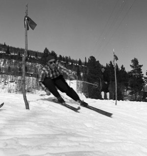 Svartvitt fotografi. En person i rutig skjorta och mörka byxor med mössa och solglasögon åker slalom. Personen är mitt i en sväng och lutar, det syns snö och markeringar i backen och några personer i bakgrunden som är på väg till backen.