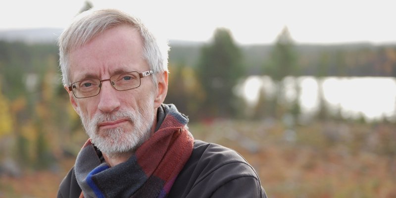 Porträttfotografi på Åke Lundgren. Åke står utomhus med en brun jack och en randig halsduk. Han har kortklippt grått hår och glasögon och ser in i kameran. Bakom honom syns ett suddigt höstlandskap med vatten, och skog.