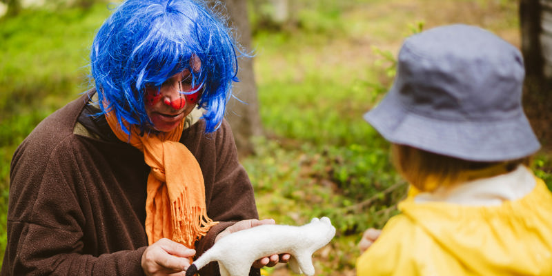 En person utklädd till figuren Plupp håller i ett tovat djur. Personen har blå peruk, orange halsduk och en brun särk i kläde på sig. I förgrunden syns ett barn med ryggen mot kameran. Bilden är tagen utomhus med ett grönskande skogsgolv runtom.