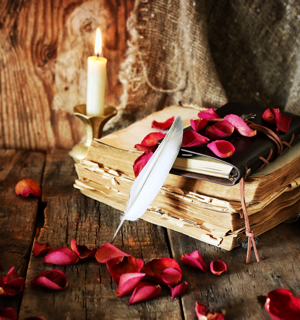 Fotografi av en trave gamla böcker som ligger på ett slitet gammalt träbord. Det står ett ljus i en ljusskake bredvid böckerna och ligger en läderinbunden skrivbok på dem. En fjäder står lutat mot traven och det ligger rosenblad utspridda över bordet och böckerna. 