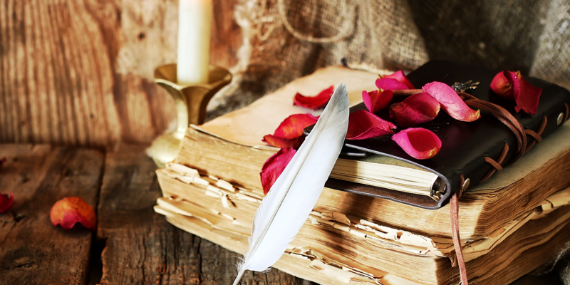 Fotografi av en trave gamla böcker som ligger på ett slitet gammalt träbord. Det står ett ljus i en ljusskake bredvid böckerna och ligger en läderinbunden skrivbok på dem. En fjäder står lutat mot traven och det ligger rosenblad utspridda över bordet och böckerna. 