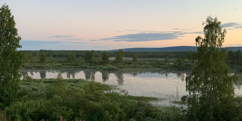 Fotografi av ett landskap med grönska, vatten och i fjärran berg. Det är skymningsljus över bilden och på himmelen syns några strimmor gråa moln. 