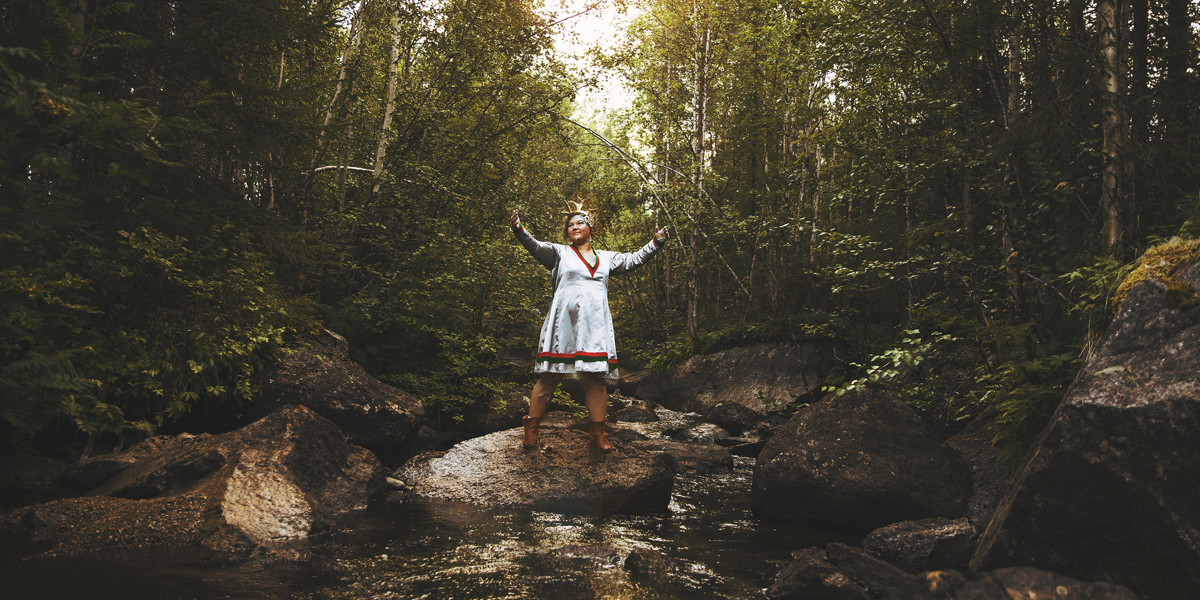 En kvinna i samisk dräkt står med händerna utsträckta på en sten i ett vattendrag.