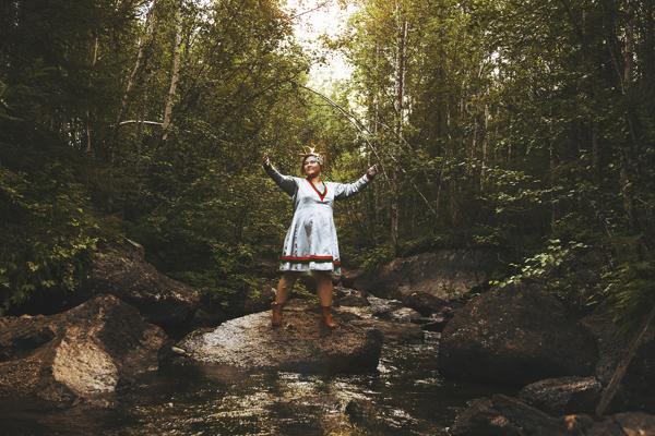 En kvinna i samisk dräkt står med händerna utsträckta på en sten i ett vattendrag.