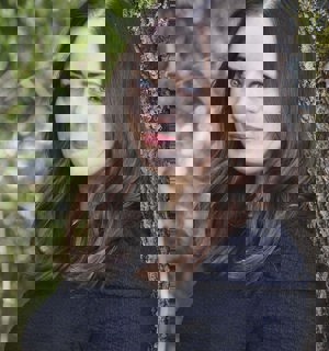 Fotografi av Stina Rundqvist. Hon står lutad mot en trästam och har en stickad tröja på sig. Hon ler med stängd mun och tittar in i kameran