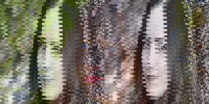 Fotografi av Stina Rundqvist. Hon står lutad mot en trästam och har en stickad tröja på sig. Hon ler med stängd mun och tittar in i kameran